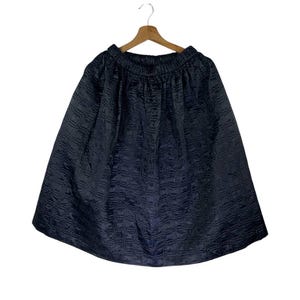 Puede incluir: Falda azul marino oscuro con un patrón acolchado texturizado. La falda está colgada de una percha de madera sobre un fondo blanco. Tiene una cintura elástica y una silueta acampanada.