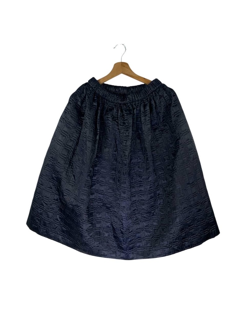 May include: A navy blue skirt with a textured, quilted design. The skirt is suspended from a wooden hanger against a white backdrop. It features an elasticated waistband and a flared shape.