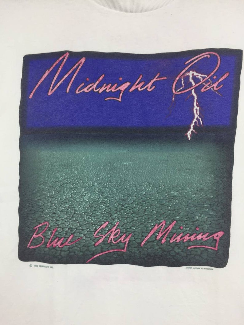 May include: White t-shirt with a graphic print of a blue sky with a lightning bolt and the text "Midnight Oil" in pink. The bottom of the graphic has a green, textured surface with the text "Blue Sky Mining" in pink.