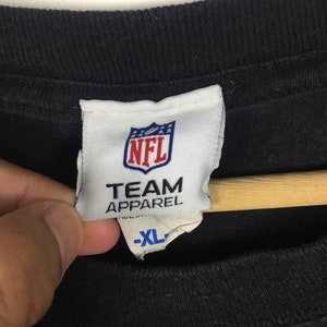 K&ouml;nnte beinhalten: Ein wei&szlig;es Etikett mit dem NFL-Logo und dem Text "TEAM APPAREL" in Blau. Das Etikett hat auch ein Gr&ouml;&szlig;enetikett mit der Aufschrift "XL" in Blau.
