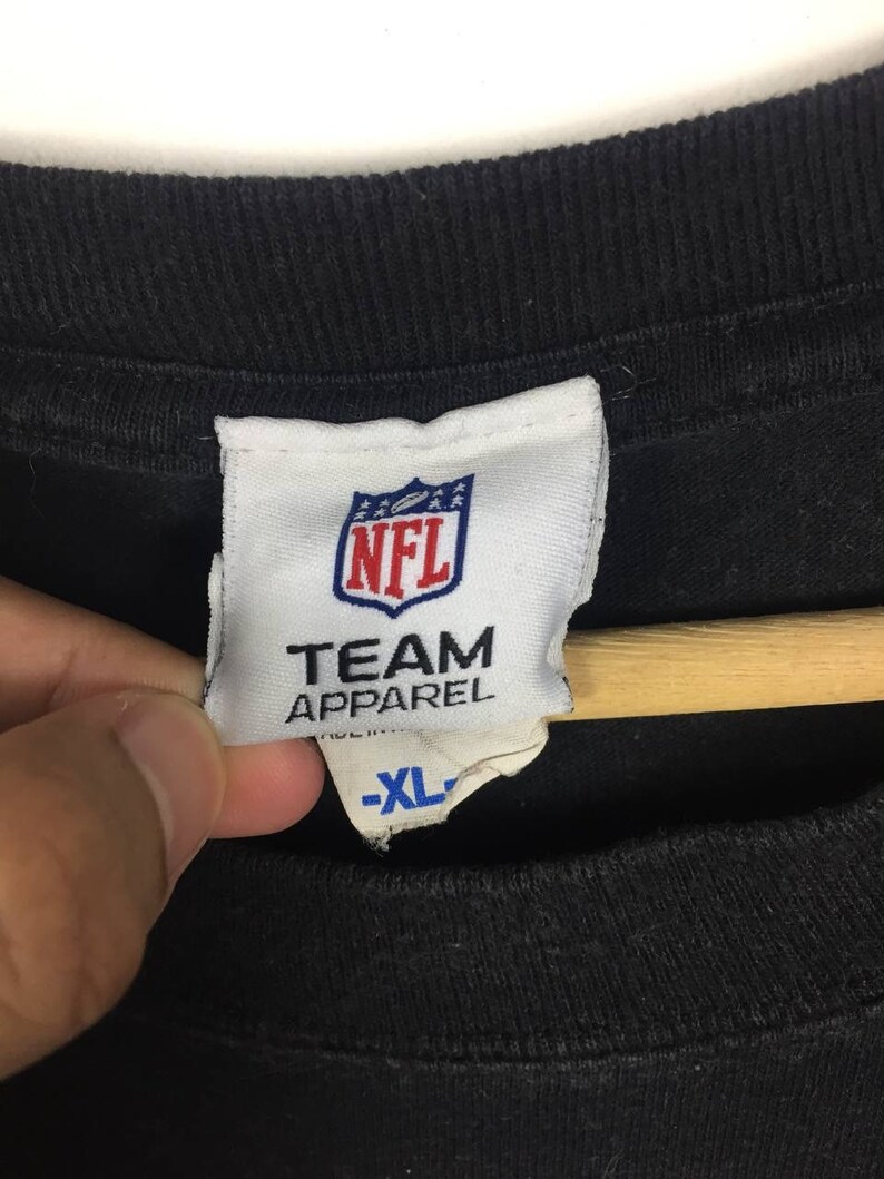 K&ouml;nnte beinhalten: Ein wei&szlig;es Etikett mit dem NFL-Logo und dem Text "TEAM APPAREL" in Blau. Das Etikett hat auch ein Gr&ouml;&szlig;enetikett mit der Aufschrift "XL" in Blau.