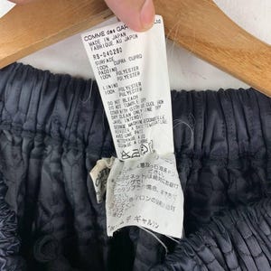 May include: A close-up of a garment tag held by a hand, with the text "COMME des GARCONS MADE IN JAPAN". The garment is a dark, quilted fabric. The tag details fabric composition and care instructions.