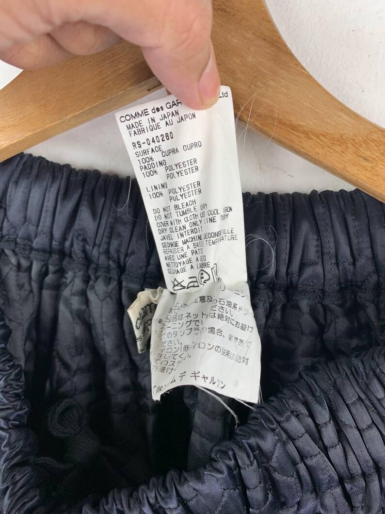 May include: A close-up of a garment tag held by a hand, with the text "COMME des GARCONS MADE IN JAPAN". The garment is a dark, quilted fabric. The tag details fabric composition and care instructions.