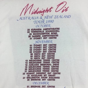May include: A white t-shirt with a pink and purple print of the Midnight Oil band tour dates for Australia and New Zealand in 1990. The tour dates are listed for October, November, and December.