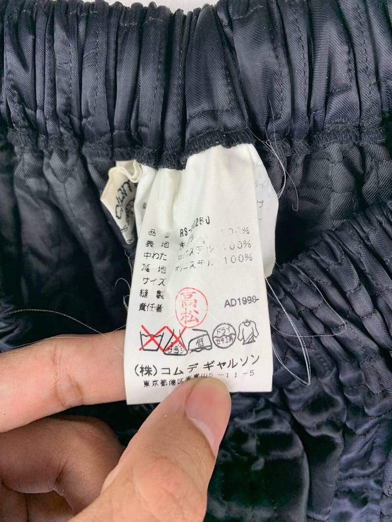 May include: Close-up of black fabric with a tag. The tag has Japanese text and symbols, including "RS-1280" and "AD1998-". The fabric appears to be quilted or textured, with a slightly shiny finish. The image is well-lit, showing the details of the fabric and tag.