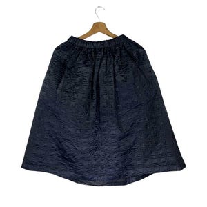 May include: A dark navy blue skirt with a textured, quilted pattern. The skirt is hanging on a wooden hanger against a white background. The skirt has an elasticated waistband and a flared silhouette.