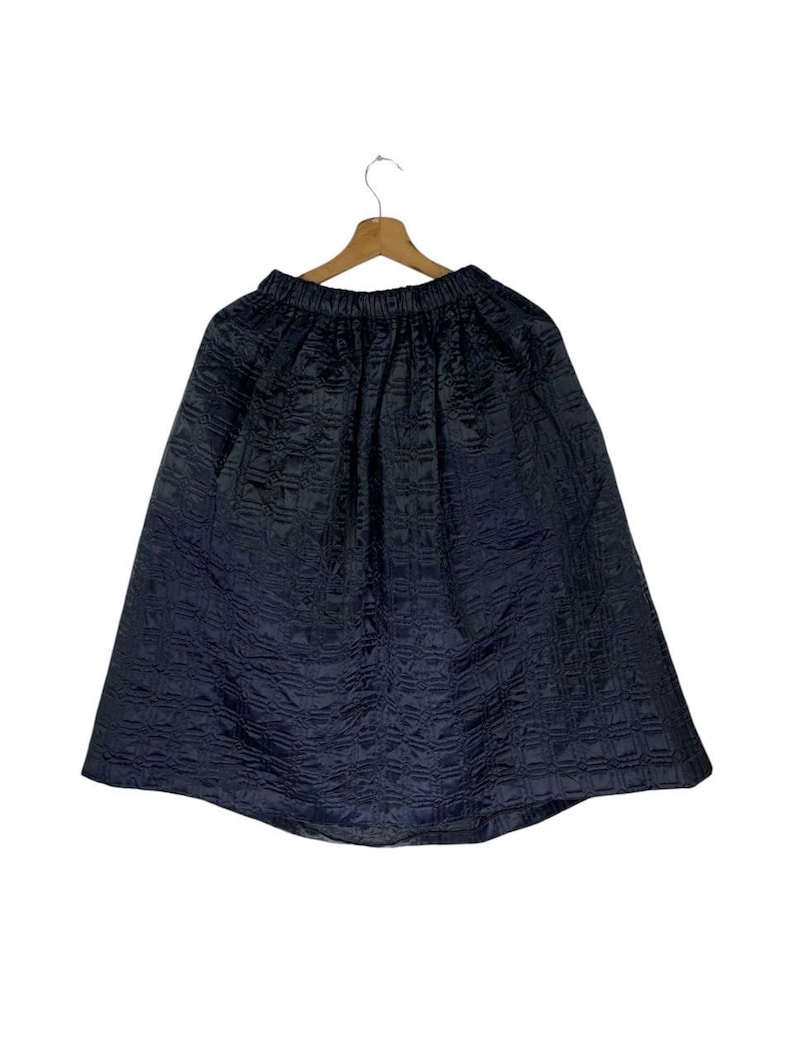 May include: A dark navy blue skirt with a textured, quilted pattern. The skirt is hanging on a wooden hanger against a white background. The skirt has an elasticated waistband and a flared silhouette.