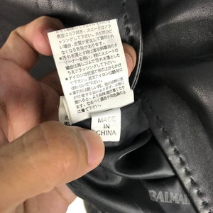 May include: Close-up of a black leather jacket care tag with instructions for cleaning and ironing. The tag is labeled "Made in China" and has the brand name "Balmain" printed on the bottom.