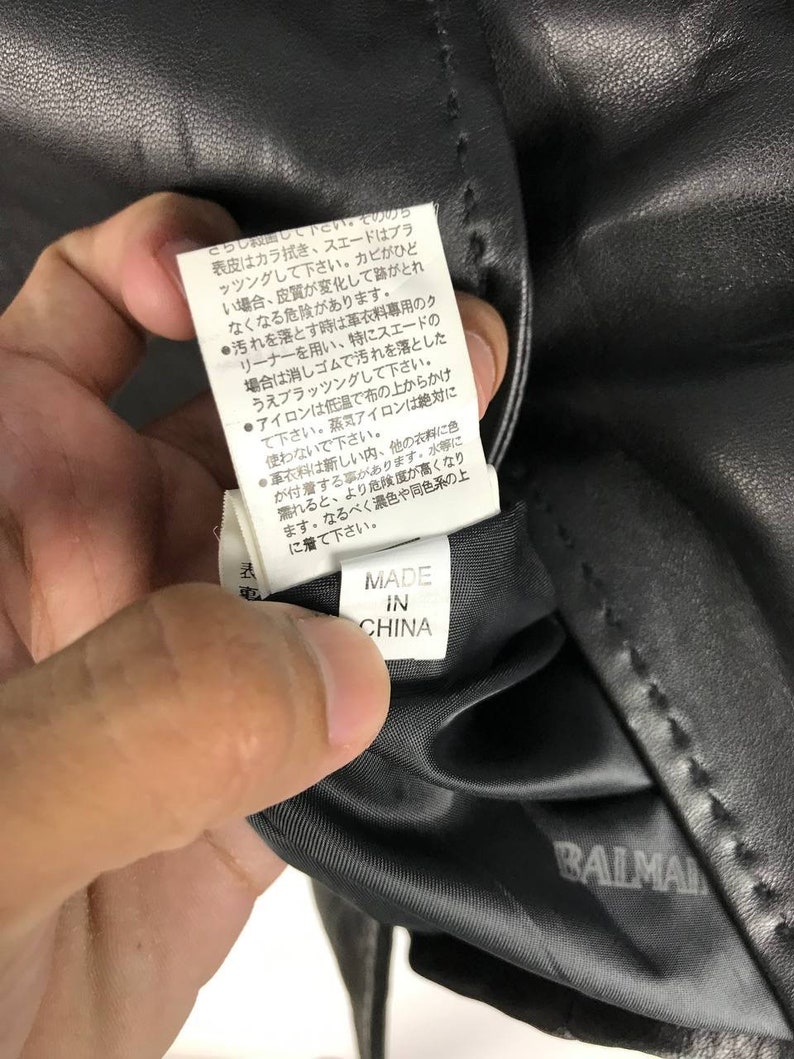 May include: Close-up of a black leather jacket care tag with instructions for cleaning and ironing. The tag is labeled "Made in China" and has the brand name "Balmain" printed on the bottom.