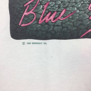 May include: White t-shirt with a black and green image of a snake skin pattern. The text "Blue" is written in pink across the image. The copyright symbol and the text "1990 MIDNIGHT OIL" are printed below the image.