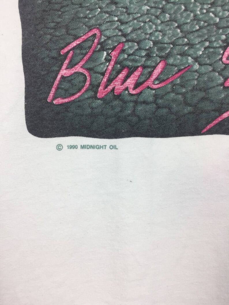 May include: White t-shirt with a black and green image of a snake skin pattern. The text "Blue" is written in pink across the image. The copyright symbol and the text "1990 MIDNIGHT OIL" are printed below the image.
