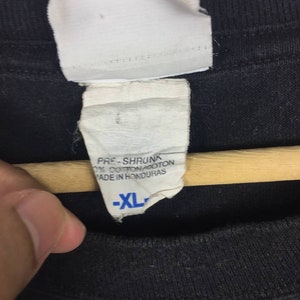 K&ouml;nnte beinhalten: Ein schwarzes T-Shirt mit einem wei&szlig;en Etikett, auf dem "Pre-Shrunk 100% Cotton Made in Honduras" und "XL" in Blau steht.