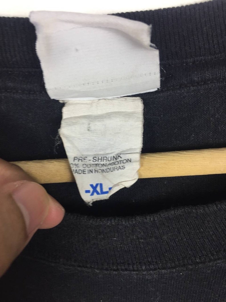 K&ouml;nnte beinhalten: Ein schwarzes T-Shirt mit einem wei&szlig;en Etikett, auf dem "Pre-Shrunk 100% Cotton Made in Honduras" und "XL" in Blau steht.