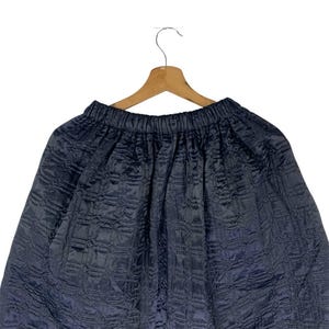 May include: A dark navy blue skirt with a quilted design, suspended from a wooden hanger. The skirt features an elasticated waistband and a flowing, voluminous silhouette. The fabric has a slight sheen.
