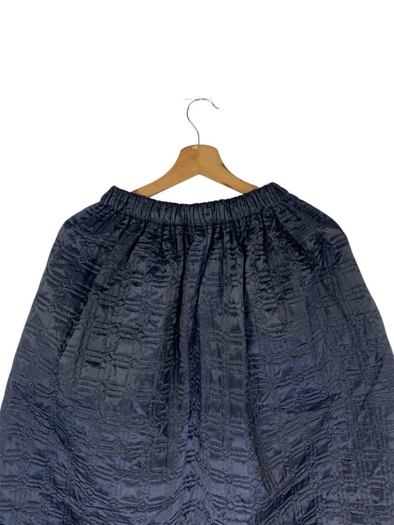 May include: A dark navy blue skirt with a quilted design, suspended from a wooden hanger. The skirt features an elasticated waistband and a flowing, voluminous silhouette. The fabric has a slight sheen.