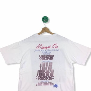 May include: White t-shirt with a pink and blue print of the Midnight Oil band tour dates for Australia and New Zealand in 1990. The text includes the dates and locations of the tour.
