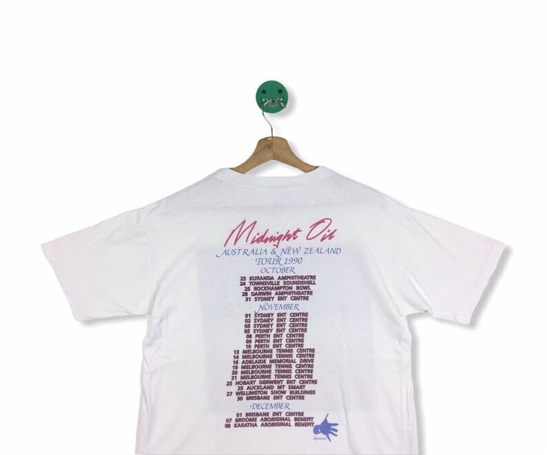 May include: White t-shirt with a pink and blue print of the Midnight Oil band tour dates for Australia and New Zealand in 1990. The text includes the dates and locations of the tour.