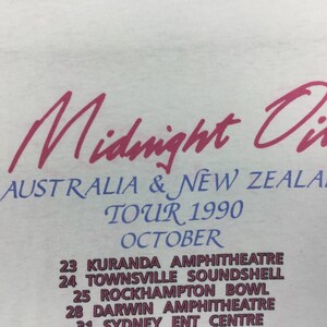 May include: A white t-shirt with a pink and blue print of the band Midnight Oil's 1990 Australia and New Zealand tour dates. The text reads "Midnight Oil", "Australia & New Zealand", "Tour 1990", "October", "Kuranda Amphitheatre", "Townsville Soundshell", "Rockhampton Bowl", "Darwin Amphitheatre", and "Sydney Ent Centre".