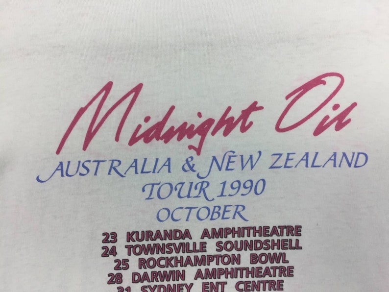 May include: A white t-shirt with a pink and blue print of the band Midnight Oil's 1990 Australia and New Zealand tour dates. The text reads "Midnight Oil", "Australia & New Zealand", "Tour 1990", "October", "Kuranda Amphitheatre", "Townsville Soundshell", "Rockhampton Bowl", "Darwin Amphitheatre", and "Sydney Ent Centre".