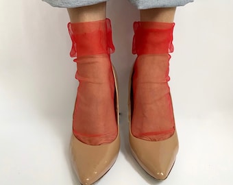 Puede incluir: Un par de calcetines de tul rojo con un borde de volantes, usados con tacones color carne y jeans azules remangados.