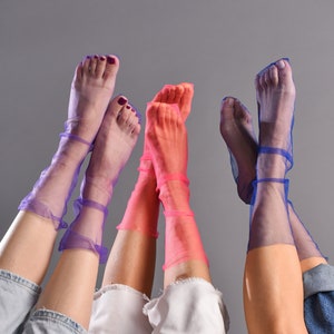 Può includere: Tre paia di gambe che indossano calze trasparenti in viola, rosa e blu. Le calze sono tutte di tonalità diverse e hanno una texture delicata e traforata. Le gambe indossano jeans e pantaloni bianchi.