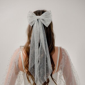 May include: A white tulle bridal hair bow with a large bow and a long trailing veil. The bow is decorated with small, sparkly embellishments.