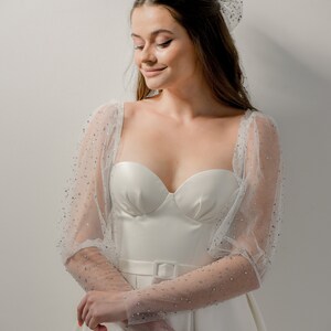 May include: A white bridal gown with a sweetheart neckline and a fitted bodice. The dress features sheer, long sleeves adorned with sparkling embellishments. A matching bow is in the hair. The dress has a belt with a square buckle.