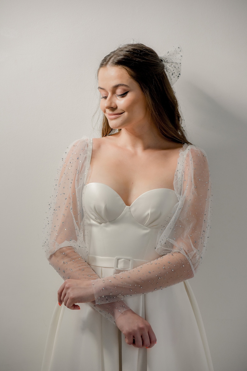 May include: A white bridal gown with a sweetheart neckline and a fitted bodice. The dress features sheer, long sleeves adorned with sparkling embellishments. A matching bow is in the hair. The dress has a belt with a square buckle.