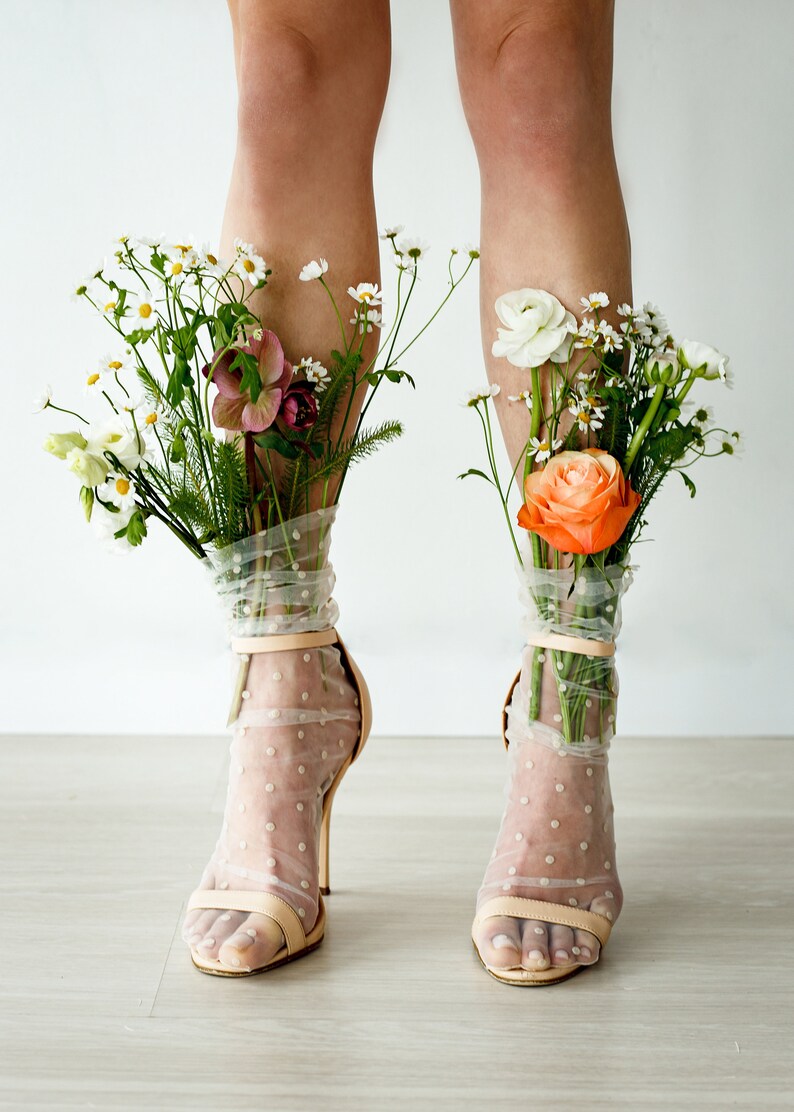 May include: A pair of nude heels with white polka dot sheer socks. The socks are filled with a bouquet of flowers, including white daisies, pink and white ranunculus, and orange roses.