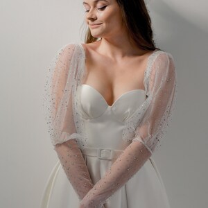 May include: A white bridal gown with a sweetheart neckline and a fitted bodice. The dress features sheer, long sleeves adorned with sparkling embellishments. A white belt cinches the waist, and the skirt flares out. A delicate hair accessory completes the look.