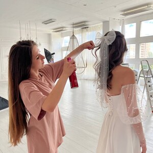 May include: A woman in a pink t-shirt and shorts is spraying hairspray on a woman wearing a white wedding dress with a large white bow in her hair.