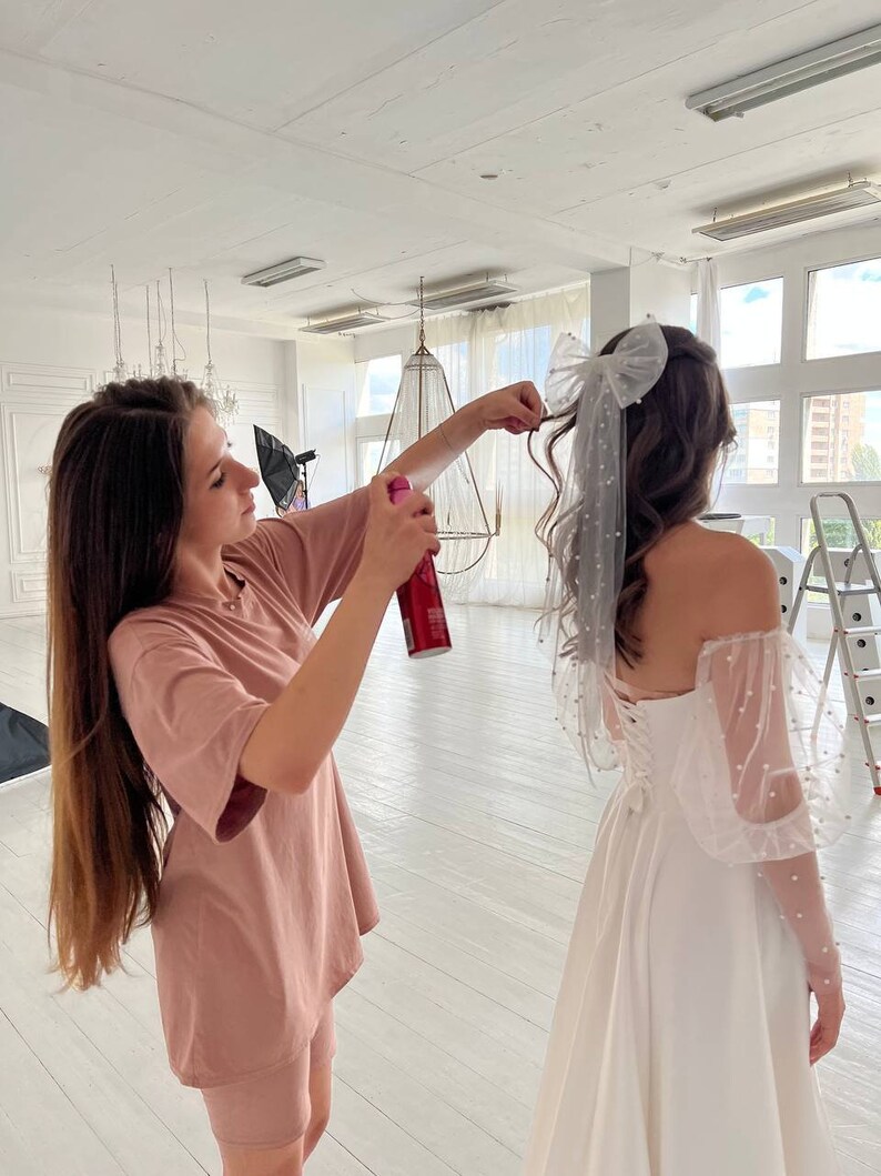 May include: A woman in a pink t-shirt and shorts is spraying hairspray on a woman wearing a white wedding dress with a large white bow in her hair.