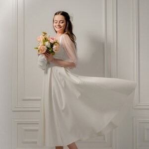 May include: A woman in a white dress with a flowing skirt and sheer, embellished sleeves, holding a bouquet of pink and peach flowers. She is wearing gold high-heeled sandals. The background is a white wall with molding.