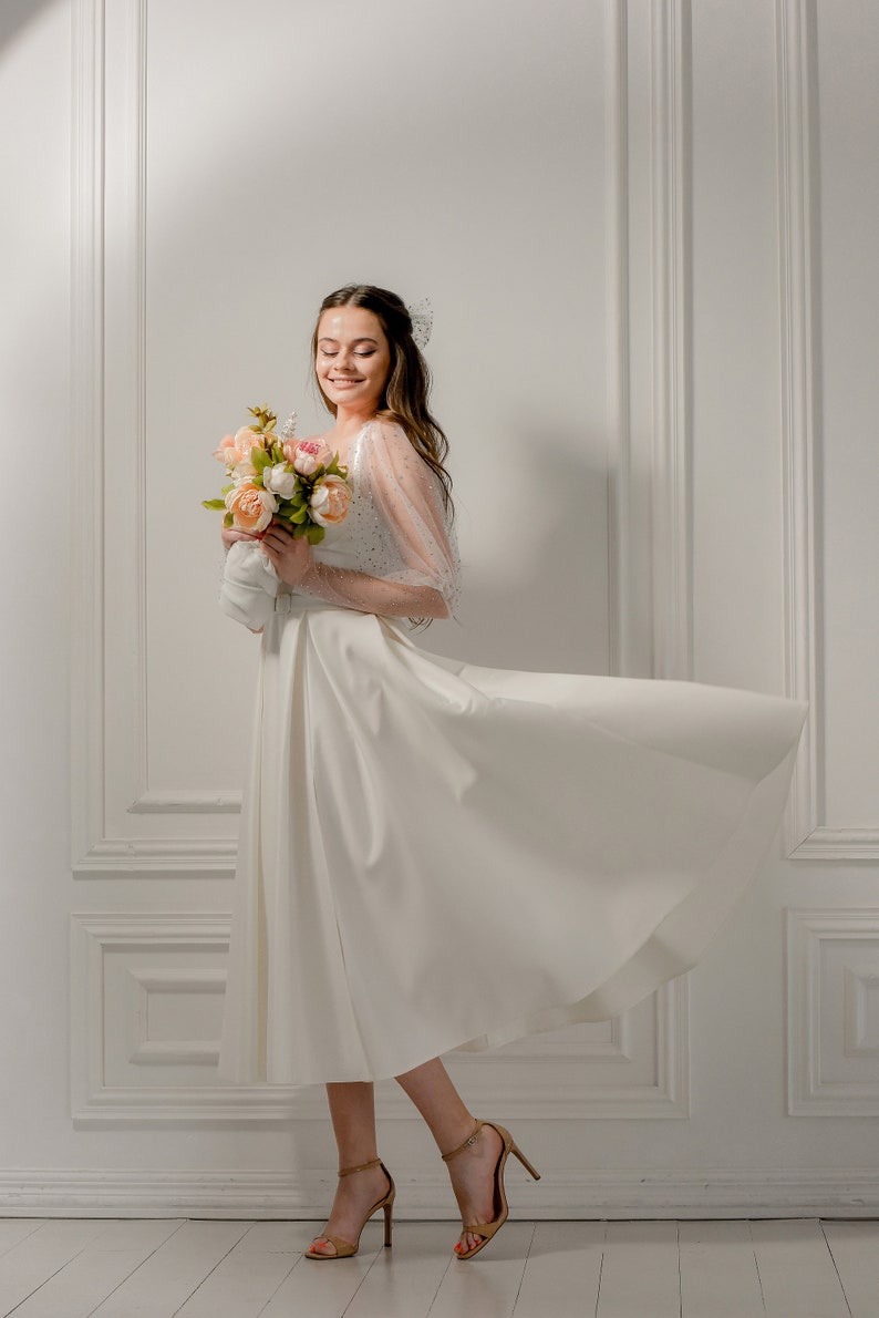 May include: A woman in a white dress with a flowing skirt and sheer, embellished sleeves, holding a bouquet of pink and peach flowers. She is wearing gold high-heeled sandals. The background is a white wall with molding.