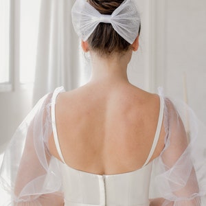 May include: A white wedding dress with a tulle overlay and a white bow in the hair.