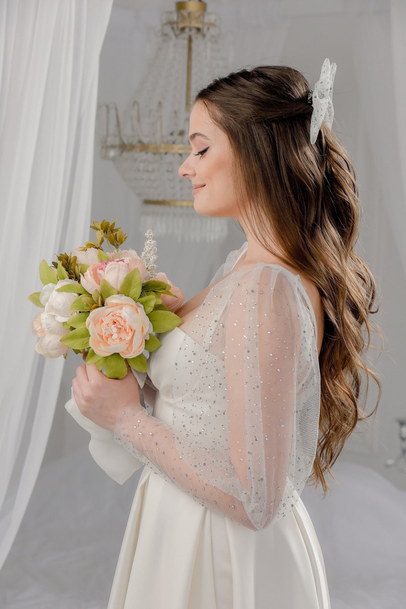 May include: A bride in a white wedding dress with sheer, sparkling sleeves holds a bouquet of peach and white flowers. A white hair accessory with rhinestones is in her hair. The background is a soft, neutral color.