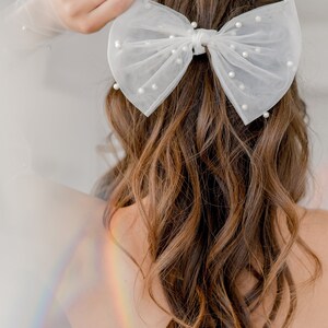 May include: A white tulle bow with pearl embellishments, worn in a woman's brown hair.