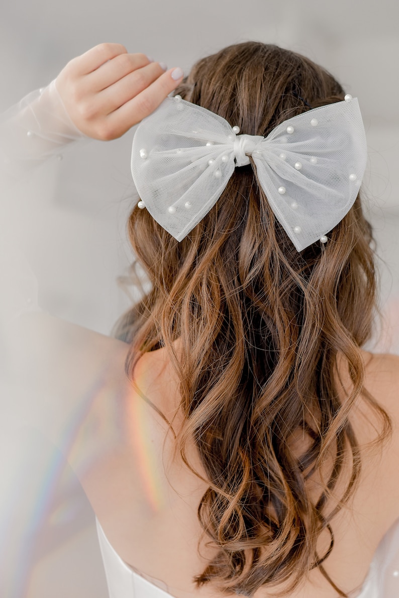 May include: A white tulle bow with pearl embellishments, worn in a woman's brown hair.