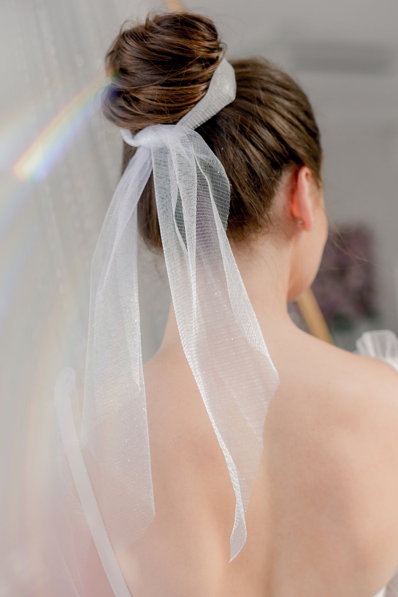 May include: A white tulle ribbon tied around a woman's brown hair bun. The ribbon is long and flowing, with a delicate, sparkly texture.