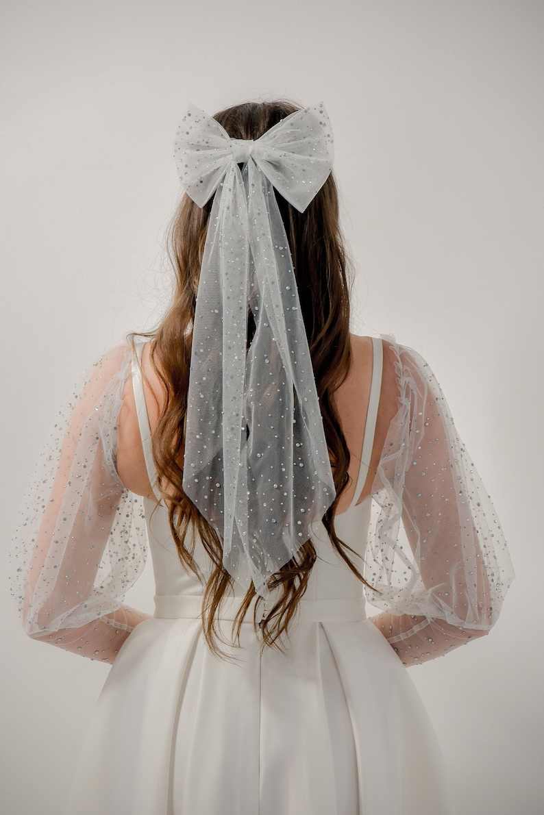 May include: A white tulle bridal hair bow with a large bow and a long trailing veil. The bow is decorated with small, sparkly embellishments.