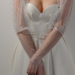 May include: A white bridal gown with a sweetheart neckline and a fitted bodice. The dress features sheer, long sleeves adorned with sparkling embellishments. A matching belt cinches the waist, creating an elegant silhouette.