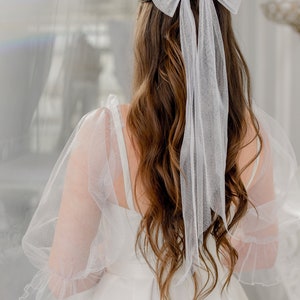 May include: A white tulle bow with long tails is tied in a woman's brown hair. She is wearing a white dress with tulle sleeves.
