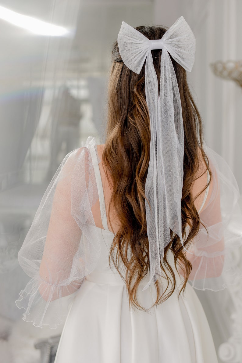 May include: A white tulle bow with long tails is tied in a woman's brown hair. She is wearing a white dress with tulle sleeves.