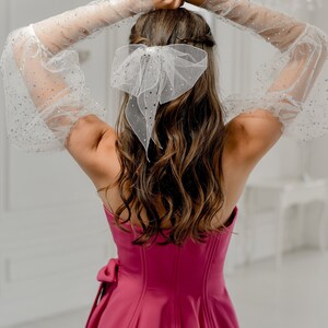 May include: A magenta-colored corset-style top with a bow detail on the side. The model is wearing sheer, long sleeves with silver embellishments and a white bow in their hair. The background is a bright, white room.