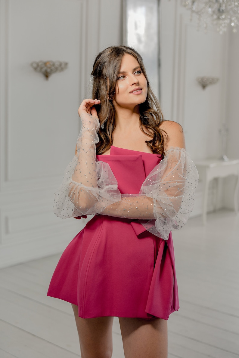 May include: A magenta-colored dress with an asymmetrical neckline and a flared skirt. The dress features sheer, long sleeves adorned with sparkling embellishments. The sleeves are puffy at the shoulders. The background is a bright, white room.