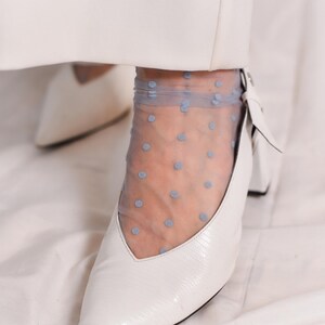May include: White pointed-toe heels with a small block heel. The shoes are worn with sheer blue polka dot socks.