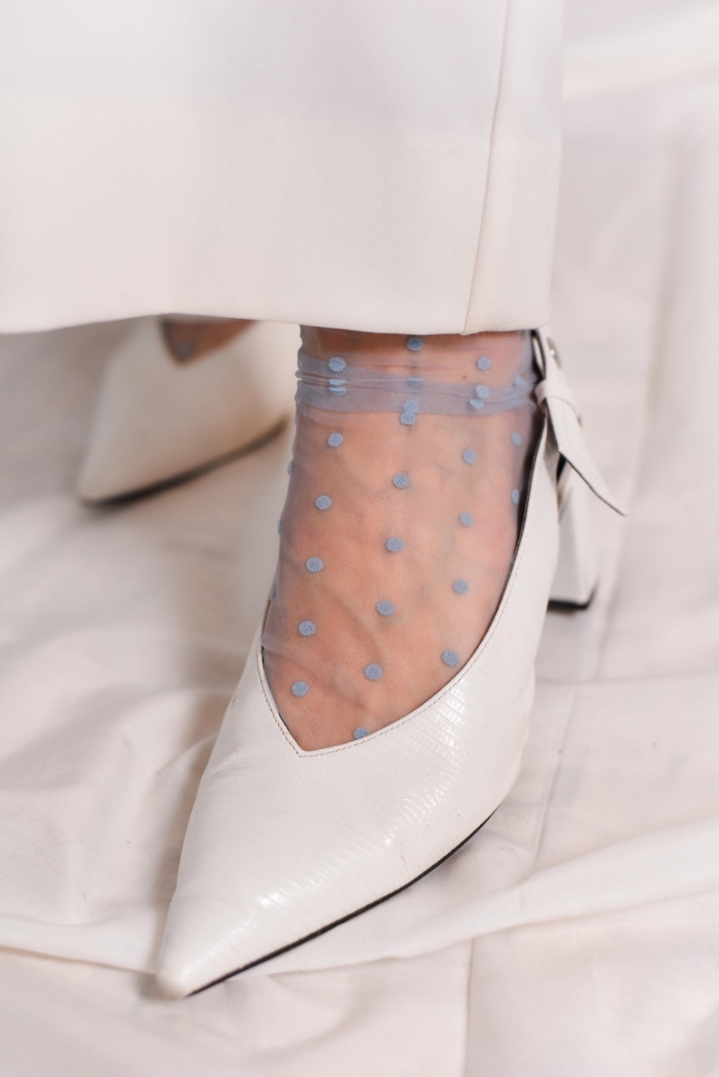 May include: White pointed-toe heels with a small block heel. The shoes are worn with sheer blue polka dot socks.