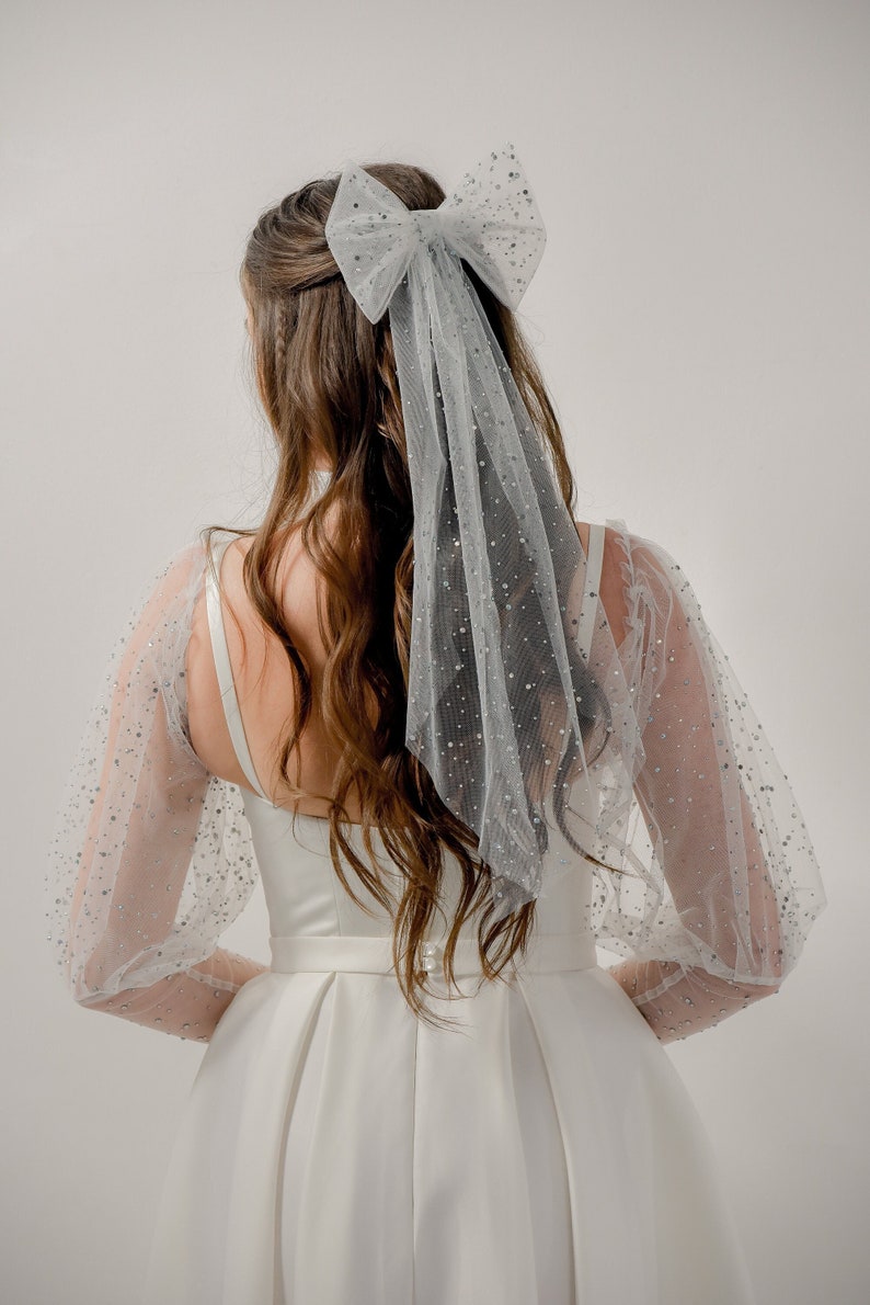 May include: A white bridal dress with a tulle veil and a sparkly bow. The dress has long sleeves with a puff design and a fitted bodice. The veil is adorned with small silver sequins.