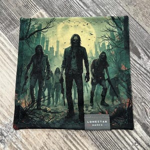 Puede incluir: Pañuelo cuadrado verde oscuro con una ilustración de zombies. La obra de arte representa varios zombis en un entorno urbano bajo la luna llena. El texto "LONESTAR HANKS" está impreso en una pequeña etiqueta gris.