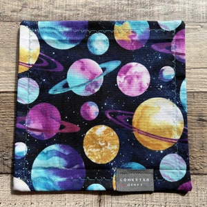 May include: Square fabric item with a space-themed pattern. The design features planets in shades of purple, blue, and gold against a dark blue background. The item has a stitched edge and a small label that reads "LONESTAR HANKS."